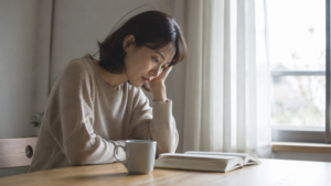 少し疲れた表情でうつむき加減。机の上にはマグカップと読みかけの本。外からやわらかな光が差し込んでいるが、女性は考え込んでいる様子